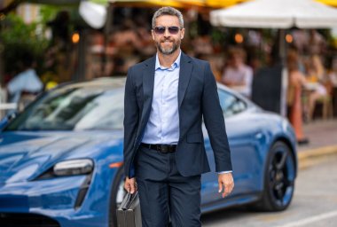 Zengin iş adamı başarısını ifade ediyor. Lüks bir iş. Pahalı arabanın yanındaki zengin adam. Lüks iş seyahati. Klasik takım elbiseli, kendine güvenen zengin bir iş adamı. Yakışıklı iş adamı evrak çantasını tutuyor..