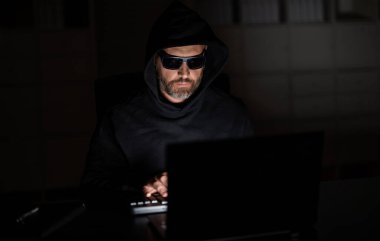 Geceleri ofiste bilgisayar korsanı. Siber saldırı. Siber suçlu ve çalıntı kişisel veriler. Dijital hırsız. Ofis laptopunda kapüşonlu bilgisayar programcısı, oyun. Hacker saldırısı. Malware enjeksiyonu.
