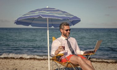 Çevrimiçi serbest çalışma. Dizüstü bilgisayarı olan bir işadamı. Kumsalda dizüstü bilgisayar kullanan bir iş adamı. Seyahat ve iş. Sea Beach 'te serbest çalışan bir adam. İş gezginleri için dinlenme yeri.