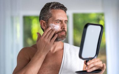 Spa adamı yüz kilinden maske takıyor. Olgun bir adam yaşlanmayı ve gençleşmeyi önlemek için nemlendirici bir maske kullanıyor. Erkek güzeli. Yaşlanma karşıtı cilt için sabah maskesi. Maske canlandırılıyor.