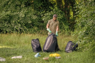 Ormanda çöp toplama. Ekoloji konsepti. Çöpleri toplayan gönüllü. Çöp torbasıyla lastik eldivenli bir adam çöpü temizliyor. Adam plastik çöp topluyor. Çevre gönüllüsü. Plastik atık sorunu.
