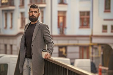 Sokakta elinde kahve tutan yakışıklı bir adam. İş hayatı, mola. Sabah kahvesi içen adam. Kahve molası. Günlük stil rutini. Sakallı adam, caddede kahve içiyor, reklam yapıyor..