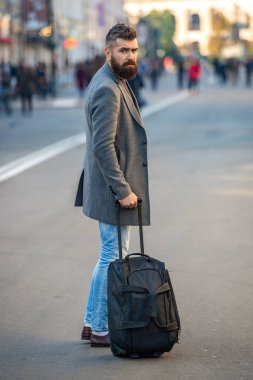 Sakallı şık bir adam şehir caddesinden seyahat çantasıyla geçip gidiyor. İş gezisi. Gezgin iş adamı. Sakallı turist şehir caddesinde yürüyor. Şehirde çantalı bir turist.