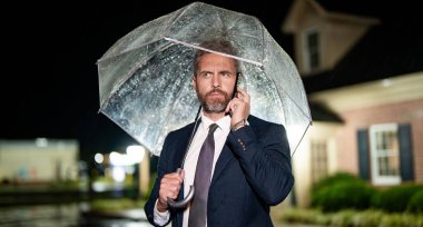 İş adamı yağmurda dikilirken telefonla konuşur. Takım elbiseli bir iş adamı cep telefonuyla konuşuyor. Yağmurlu sokakta telefonu olan bir iş adamı. Yağmurda iş adamı. Şemsiye altında iş görüşmesi. Telefon..