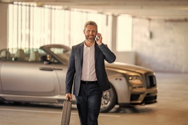 Takım elbiseli olgun bir iş adamı iş görüşmesi yapıyor. Şirket işi. Takım elbiseli zengin iş adamı. İş portresi. Başarılı bir iş adamı park yerinde evrak çantasıyla yürüyor. İş adamı evrak çantasını tutar.