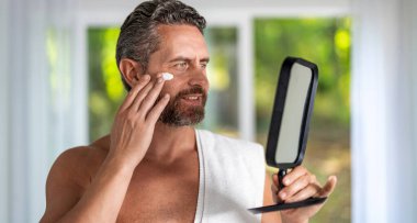 Erkek yüzü için krem. Yaşlanma önleyici ve kırışıklık önleyici krem. Adam aynaya kozmetik yüz kremi sürüyor. Deri kozmetiği. Güzellik kremi ya da yaşlanmayı önleyen cilt bakımı. Erkekler için makyaj malzemesi. Sağlıklı ışıltı.