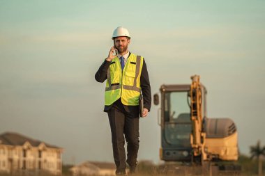 Bina sorumlusunun telefonu var. Hardhat 'ın baş mühendisi telefon kullanıyor. İş adamı telefonda konuşur. İnşaat mühendisi. Denetmen iş adamı. Şantiyedeki adam. İş projesi iletişimi.