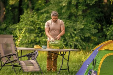 Kampta taze yiyecek. Adam yaz kampında karpuz kesiyor. Yaz günü doğa yürüyüşçüsü. Kamp ve doğa pikniği. Adam kampı. Karpuz kampı pikniği. Lezzetli sağlıklı yiyecekler. Yemek zamanı..
