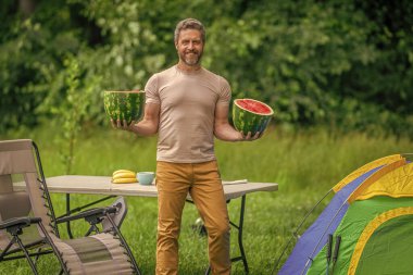 Kampta taze yiyecek. Yaz kampında karpuzlu bir adam. Yaz günü doğa yürüyüşçüsü. Kamp ve doğa pikniği. Adam kampı. Karpuz kampı pikniği. Lezzetli sağlıklı yiyecekler. Doğa geri çekiliyor..