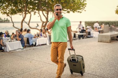 Gezgin telefon kullanıyor. Turist tatili rezervasyonu. Yaz gezisi için seyahat valizi. Yaz gezisini ayarlayan adam. İspanyol turist gezginleri kitabı internette. Seyahat çantalı bir turist. Uçuş rezervasyonu.