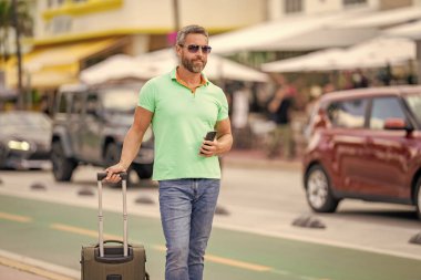 Yaz gezisi için seyahat valizi. Yaz gezisini ayarlayan adam. İspanyol turist gezginleri kitabı internette. Seyahat çantalı bir turist. Gezgin telefon kullanıyor. Turist tatili rezervasyonu. Bilet al.