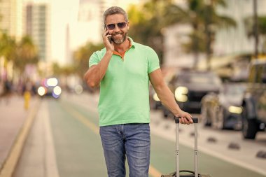 Yaz gezisini ayarlayan adam. İspanyol turist gezginleri kitabı internette. Seyahat çantalı bir turist. Gezgin telefon kullanıyor. Turist tatili rezervasyonu. Yaz gezisi için seyahat valizi. İnternetten rezervasyon.