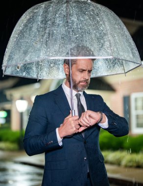 Geç ve dakik. Dakik bir iş adamı. Yağmurlu havada bekleyen bir adam. İş adamı şemsiyeyle yağmurun altında bekler. İş adamı caddede nöbet tutuyor. İş adamı kontrol zamanı. İş teslim tarihi.