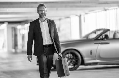 İş adamı evrak çantasını tutar. Başarılı bir anlaşmadan sonra takım elbiseli mutlu bir işadamı. Şirket işi. Takım elbiseli zengin iş adamı. İş portresi. Başarılı bir iş adamı park yerinde evrak çantasıyla yürüyor..