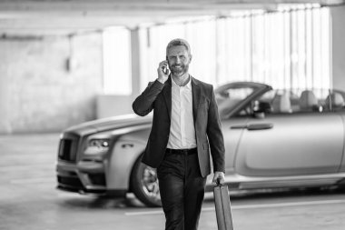 Takım elbiseli olgun bir iş adamı iş görüşmesi yapıyor. Şirket işi. Takım elbiseli zengin iş adamı. İş portresi. Başarılı bir iş adamı park yerinde evrak çantasıyla yürüyor. İş adamı evrak çantasını tutar.