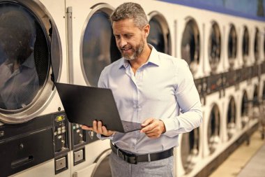 Çamaşır ve iş yükleme işinde çok çalışan var. Çamaşır ve teslim tarihleriyle uğraşan yorgun bir işçi. Yoğun iş adamları için aşırı çamaşır yüklemesi. Yoğun bir program. Çamaşırhanede dizüstü bilgisayarda çalışan meşgul bir adam..