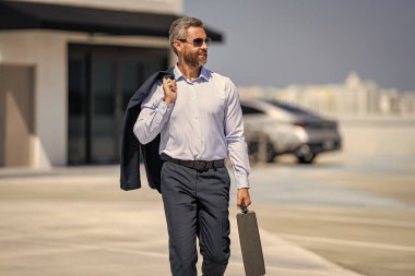 Takım elbiseli İspanyol iş adamı. İş başarısı. Başarılı bir iş adamı yürüyor. Başarılı bir iş adamı park yerinde evrak çantasıyla yürüyor. İş adamı evrak çantasını tutar. Kendine güvenen lider.