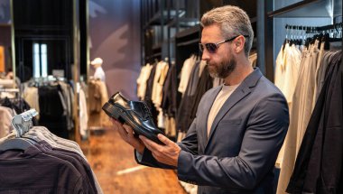 Güneş gözlüklü bir iş adamı dükkanda şık ayakkabılar seçer. Resmi görünmek için klasik deri ayakkabılar. Resmi ayakkabı. Deri ayakkabılar. Ayakkabı mağazasındaki tezgahtar adam klasik ayakkabıları tutuyor. Ayakkabı mağazası.