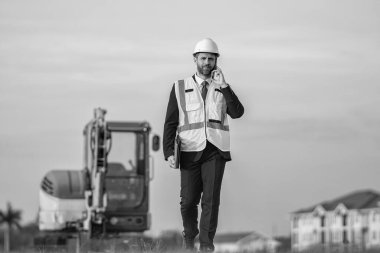 Bina sorumlusunun telefonu var. Hardhat 'ın baş mühendisi telefon kullanıyor. İş adamı telefonda konuşur. İnşaat mühendisi. Denetmen iş adamı. Şantiyedeki adam. İş projesi iletişimi.