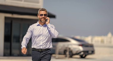 Başarılı bir iş adamı otoparkta yürüyor. Takım elbiseli olgun bir iş adamı iş görüşmesi yapıyor. Şirket işi. Takım elbiseli zengin iş adamı. İş portresi reklamı.