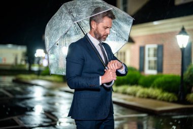 İş adamı kontrol zamanı. Geç ve dakik. Dakik bir iş adamı. Yağmurlu havayı bekleyen bir adam. İş adamı şemsiyeyle yağmurun altında bekler. İş adamı caddede nöbet tutuyor. Görüşmeyi bekliyorum..