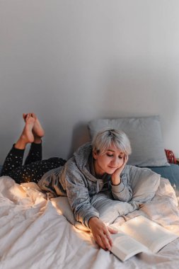 Sarı saçlı genç bir kadın yatakta kitap okuyor. Sağlıklılık. Dijital detoks. Hayatımızın fişi çekiliyor. Pijama takımı. Sıcak bir tatil günü. Noel ışıkları. Rahat giysiler. Gri, bej, beyaz.