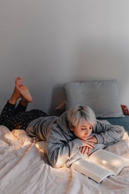 Sarı saçlı genç bir kadın yatakta kitap okuyor. Sağlıklılık. Dijital detoks. Hayatımızın fişi çekiliyor. Pijama takımı. Sıcak bir tatil günü. Noel ışıkları. Rahat giysiler. Gri, bej, beyaz.