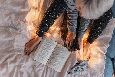 Sarı saçlı genç bir kadın yatakta kitap okuyor. Sağlıklılık. Dijital detoks. Hayatımızın fişi çekiliyor. Pijama takımı. Sıcak bir tatil günü. Noel ışıkları. Rahat giysiler. Sevimli kedicik..