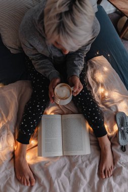 Sarı saçlı genç bir kadın yatakta kitap okuyor. Sağlıklılık. Dijital detoks. Hayatımızın fişi çekiliyor. Pijama takımı. Sıcak bir tatil günü. Noel ışıkları. Rahat giysiler. Sıcak kahve içiyorum..