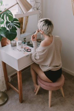 Genç milenyum kadını giyiniyor. Küpe takıyordu. Güzellik rutini. Makyöz. Maskara, ruj, allık. Pastel renkler. Makyaj aynası. Güzel sarışın bayan. Beyaz yatak odası ışığı.