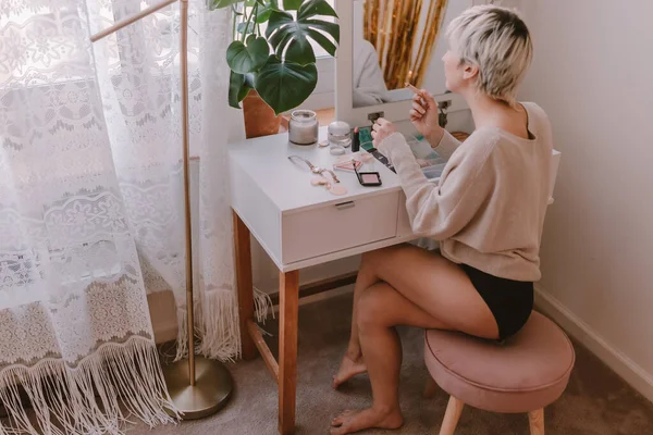 Young millennial woman is dressing up. Wearing earrings. Beauty routine. Make up artist. Mascara, lipstick, blushes. Pastel colors. Dressing vanity mirror. Pretty blonde lady. White bedroom light.