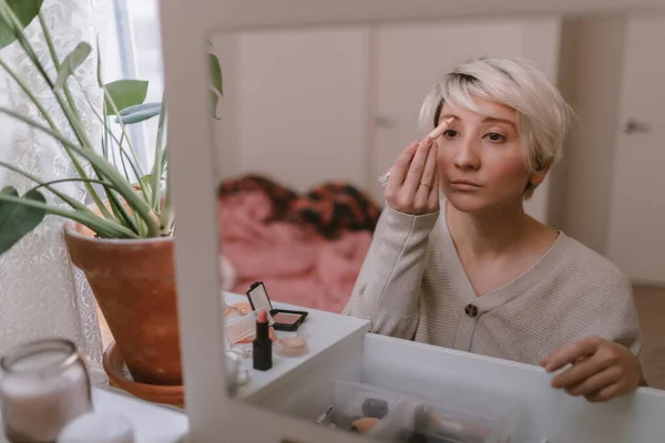 Young millennial woman is dressing up. Wearing earrings. Beauty routine. Make up artist. Mascara, lipstick, blushes. Pastel colors. Dressing vanity mirror. Pretty blonde lady. White bedroom light.