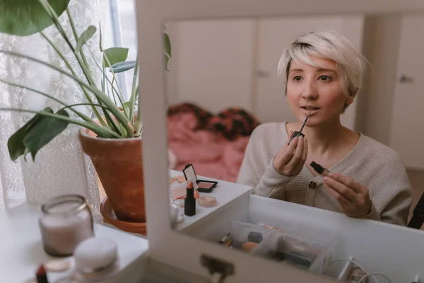 Young millennial woman is dressing up. Wearing earrings. Beauty routine. Make up artist. Mascara, lipstick, blushes. Pastel colors. Dressing vanity mirror. Pretty blonde lady. White bedroom light.