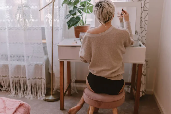 Young millennial woman is dressing up. Wearing earrings. Beauty routine. Make up artist. Mascara, lipstick, blushes. Pastel colors. Dressing vanity mirror. Pretty blonde lady. White bedroom light.