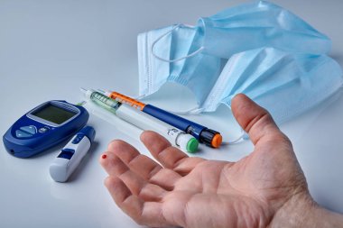 Parmağında bir damla kan, insülin şırınga kalemleri ve tıbbi maskelerle.