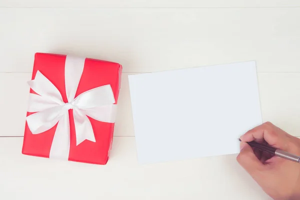 空白のグリーティングカードと木製のテーブル プレゼントやパッケージ ポストカードモックアップ トップビュー フラットレイ コピースペース 休日のコンセプトに赤いギフトボックスを持つ男の手紙を書くの手 — ストック写真