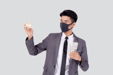 Yüz maskeli genç Asyalı işadamı para dolarını elinde tutuyor ve beyaz arka planda, salgın hastalıklar sırasında ekonomik, iş adamı ve yatırım mülkünde kriz ve izole edilmiş bir evde yaşıyor..