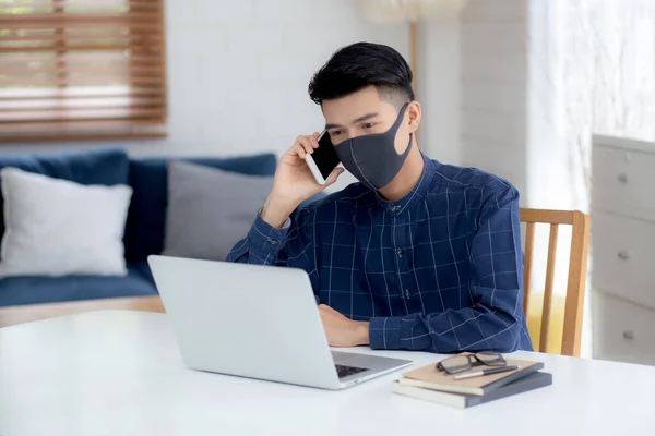 顔のマスクの若いアジアのビジネスマンは スマートフォンを話し ノートパソコン上で動作するCovid 19を保護するために ビジネスマンの隔離作業ノートブック 社会的距離と自宅から家に滞在 — ストック写真