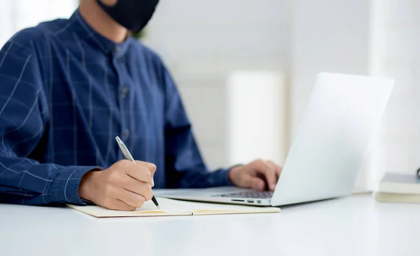Young asian business man in face mask for protect covid-19 and work on laptop computer and writing notebook, businessman quarantine work from home, new normal, social distancing and stay home.