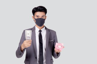 Yüz maskeli genç Asyalı işadamı elinde dolar ve domuz kumbarası ile beyaz arka planda izole edilmiş, ekonomik kriz, maske yatırımcısı finans ve hastalık sırasında tasarruf yapan iş adamı.