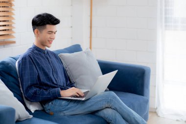 Genç Asyalı iş adamı evde dizüstü bilgisayarla gülüyor ve çalışıyor oturma odasındaki kanepede internete bağlanıyor, serbest çalışan adam kanepeye dizüstü bilgisayar kullanıyor, konforlu, yeni normal yaşam tarzı konsepti..