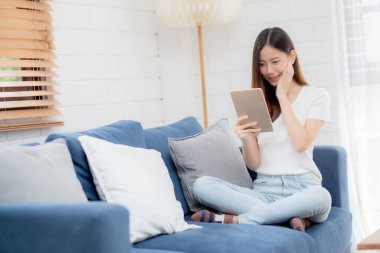 Çekici Asyalı bir kadın evdeki koltukta bilgisayarını kullanarak dinleniyor, mutlu bir kız kanepeye oturmuş heyecanlı, iletişim ve yaşam tarzı konsepti olan dijital aletleri okuyor..