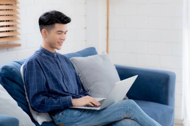 Genç Asyalı iş adamı evde dizüstü bilgisayarla gülüyor ve çalışıyor oturma odasındaki kanepede internete bağlanıyor, serbest çalışan adam kanepeye dizüstü bilgisayar kullanıyor, konforlu, yeni normal yaşam tarzı konsepti..