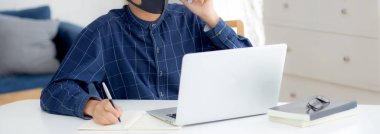 Yüz maskeli Asyalı genç bir iş adamı akıllı telefondan konuşuyor ve bilgisayarla covid-19 'u koruyor, iş adamı karantina işini evden yapıyor ve not defteri yazıyor, sosyal mesafe koyuyor ve evde kalıyor..