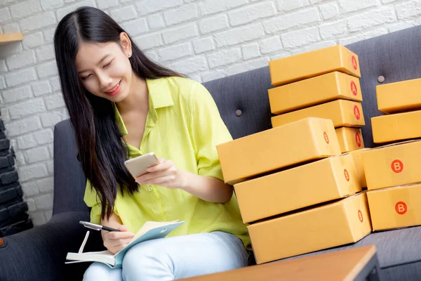 年轻的亚洲女人是个企业家 在家里用笔记本电脑上网 网上购物 物流和送货 看客户的电话和书写订单 女售货员在沙发上工作 女商人是中小企业 — 图库照片