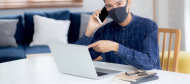 Yüz maskeli Asyalı genç bir iş adamı akıllı telefondan konuşuyor ve bilgisayar başında Coperid-19 'u korumak için çalışıyor. İş adamı karantina işini evden defter ve sosyal mesafeyle yapıyor ve evde kalıyor..