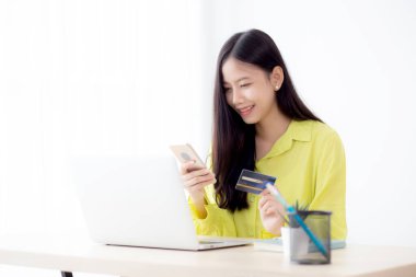 Genç Asyalı iş kadını akıllı telefon kullanıyor ve kredi kartını tutarken çevrimiçi alışveriş ve ödemeyi evdeki dizüstü bilgisayarla yapıyor, kadın banka kartı tutuyor, iletişim konsepti.