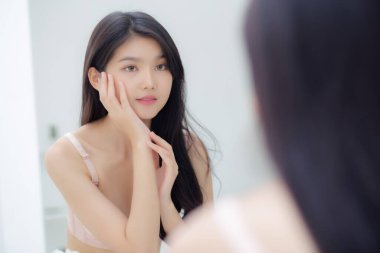 Genç ve güzel Asyalı bir kadın gülümsüyor cilt bakımı ve kozmetik ile cilt bakımını kontrol ederken aynaya bakıyor. Güzellik kızı, krem ve losyonla mutlu bir yüz ifadesi sergiliyor..