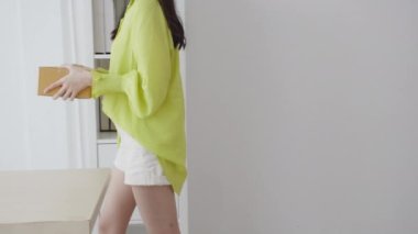 Genç Asyalı bir kadın elinde karton kutuyla eve gidiyor, mutlu bir kadın koli kutusu taşıyor ve onu masanın üstüne koyup hediye paketi, hediye paketi, online alışveriş mağazası ve servis konseptine bakıyor..