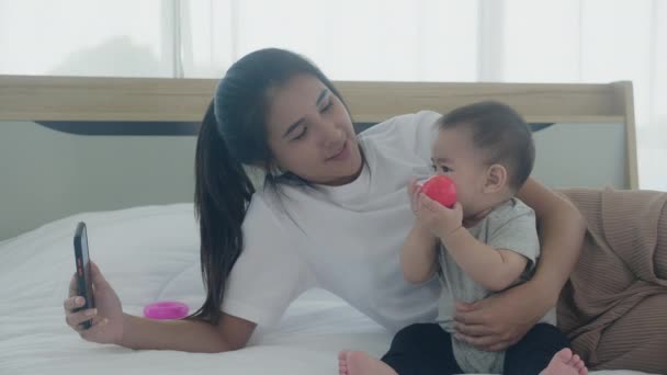 若いですアジアの母親と小さな赤ちゃんの女の子または新生児自画とともにベッドの上でスマートフォンでベッド 幸福の母と娘使用して電話ビデオ通話で自宅 家族と通信コンセプト — ストック動画